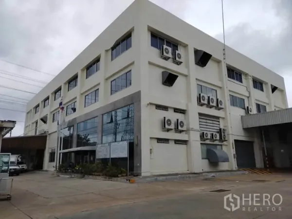 Factory for rent or sale in Bang Sao Thong, Samut Prakan - Multi-storey factory and office building with large glass frontage, exterior air-conditioning units, loading bay and roller shutter, set on a paved yard.
