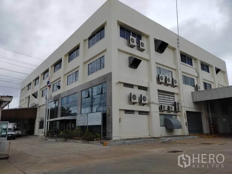 Factory for rent or sale in Bang Sao Thong, Samut Prakan - Multi-storey factory and office building with large glass frontage, exterior air-conditioning units, loading bay and roller shutter, set on a paved yard.