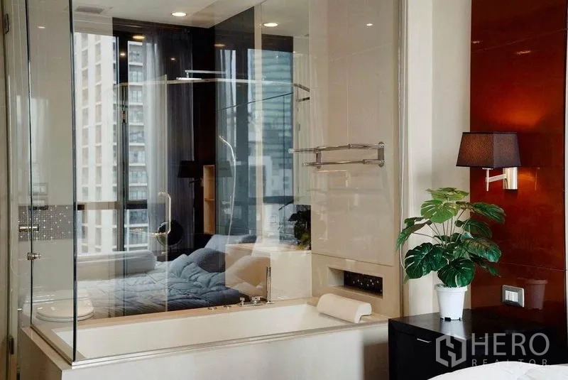 Condominium for rent in Khlong Toei, Bangkok - Bathtub next to glass wall with bedside lamp and potted plant, high‑rise city view.