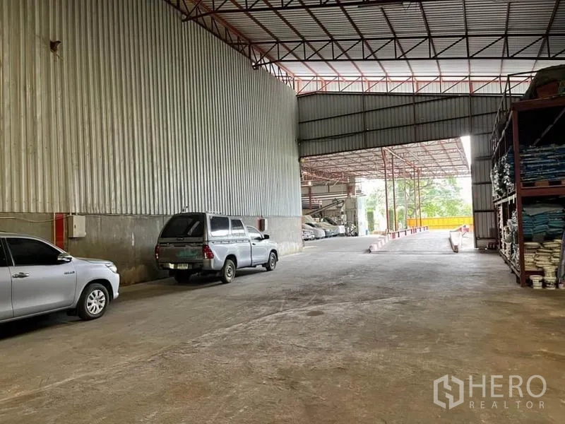 Warehouse for sale in Lam Luk Ka, Pathum Thani - Interior passage opening to the yard, with racking on the right and vehicles along the wall.