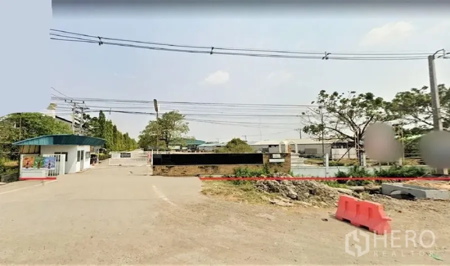 Factory for sale in Lam Luk Ka, Pathum Thani - Gated industrial entrance in Lam Luk Ka with security booth, boundary wall and wide access road under power lines.