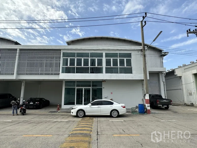 工厂 出租在Khlong Luang, Pathum Thani - 工厂外立面，双层玻璃办公室、卷帘门与停车位，园区内道路。