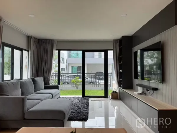 Detached House for rent in Bang Phli, Samut Prakan - Bright living room with grey sectional sofa, TV wall and sliding doors to the garden.