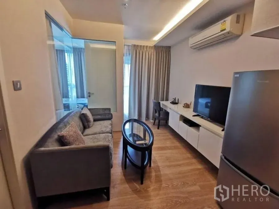Condominium for rent or sale in Watthana, Bangkok - Living room with gray sofa, glass coffee table, TV and work desk by large windows.