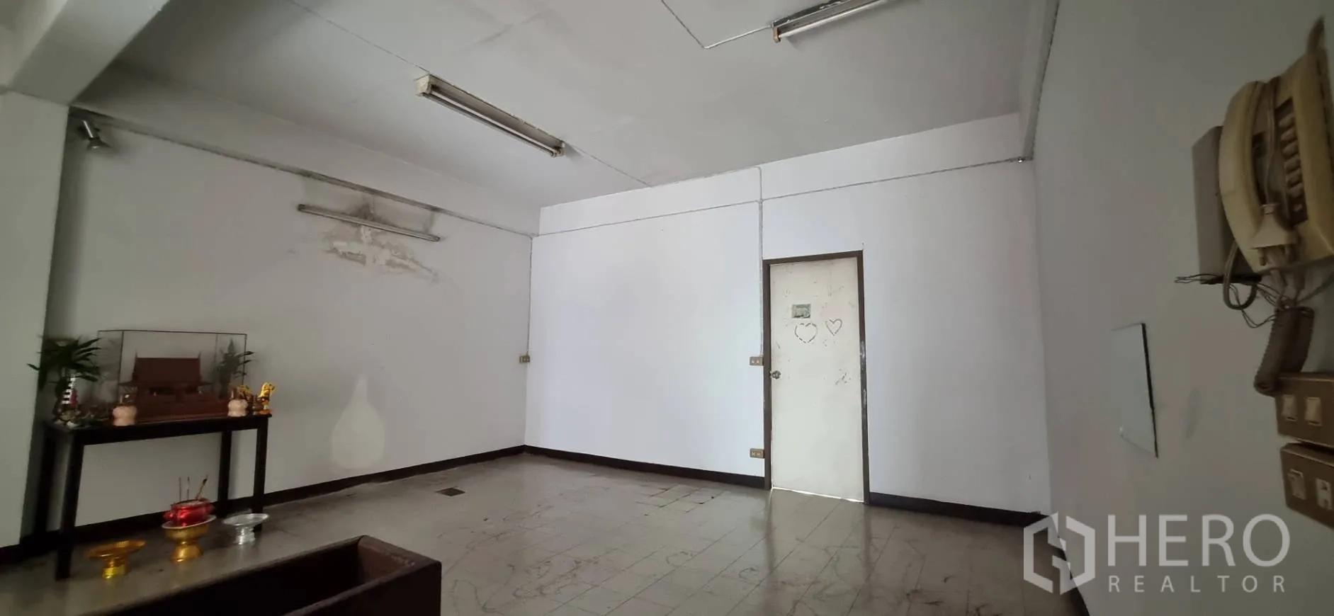 Shophouse for rent or sale in Bangkok - Simple room with tiled floor and a wall-mounted shrine table under fluorescent lights.