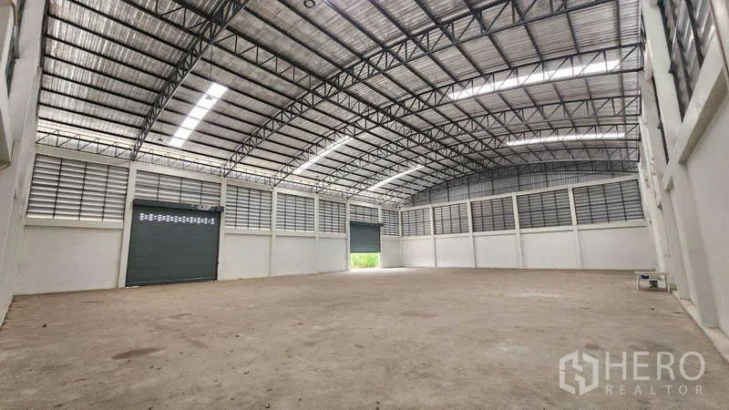 Factory for rent in Ban Phaeo, Samut Sakhon - Spacious factory interior with high arched steel roof, skylight strips, louvered vents and a large roller shutter door.