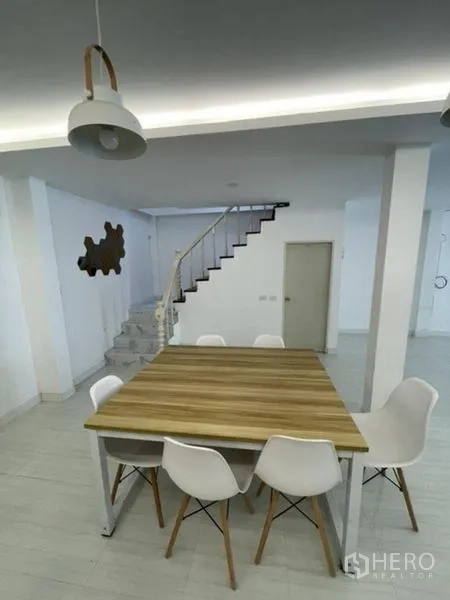 Townhouse for rent in Phra Khanong, Bangkok - Dining area with a wood table beneath pendant lights and stairs to upper floors.