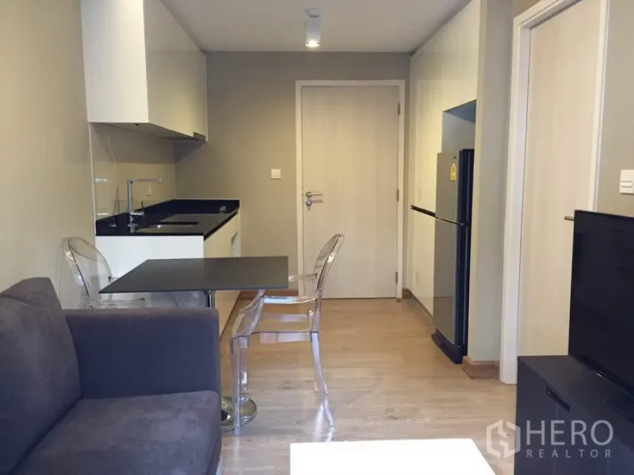 Condominium for rent in Watthana, Bangkok - Compact living and dining area with kitchenette, black countertop, and clear chairs in a modern Bangkok condo.