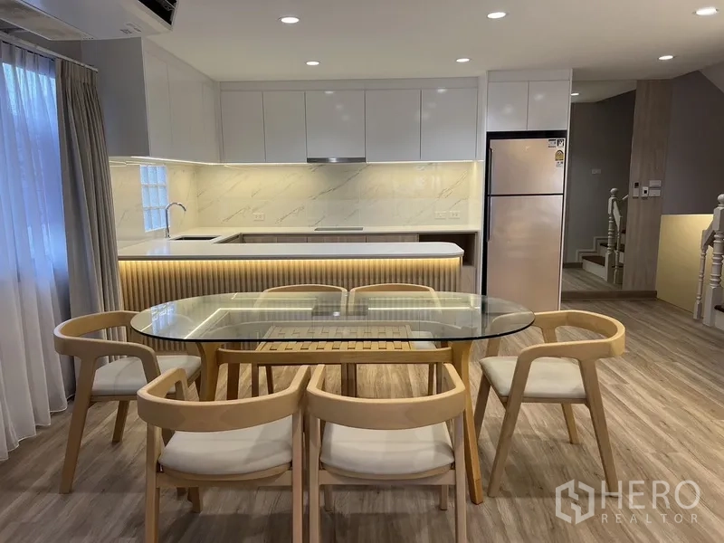 Detached House for rent in Bang Rak, Bangkok - Glass dining table for six beside an illuminated white marble-look kitchen with island and stainless fridge.