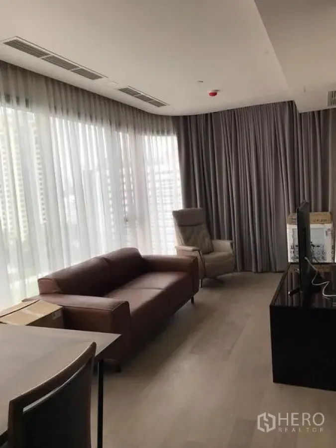 公寓 出租在Watthana, Bangkok - 客厅采光充足，落地窗与纱帘，棕色皮沙发与电视柜，可望城市景观。