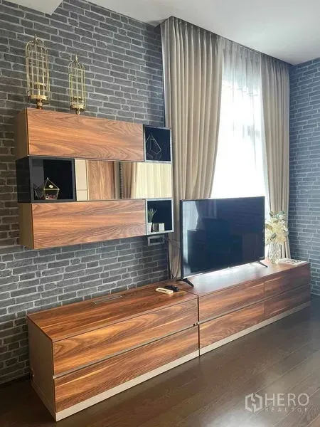 Condominium for rent or sale in Watthana, Bangkok - Living area with brick accent wall, wood media cabinets and flat-screen TV beside tall curtains.
