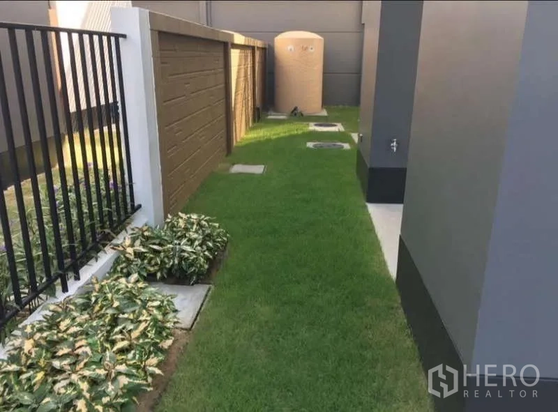 Detached House for rent in Bang Kapi, Bangkok - Narrow side yard with green grass, utility tank and planted borders along the fences.