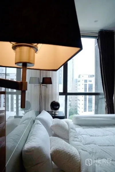 Condominium for rent in Khlong Toei, Bangkok - Cozy bedroom with white bedding, bedside lamps, and floor‑to‑ceiling window overlooking the skyline.