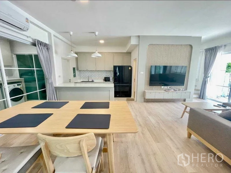 Townhouse for rent or sale in Prawet, Bangkok - Open living space showing dining table, pendant lights, kitchen and a feature TV wall.