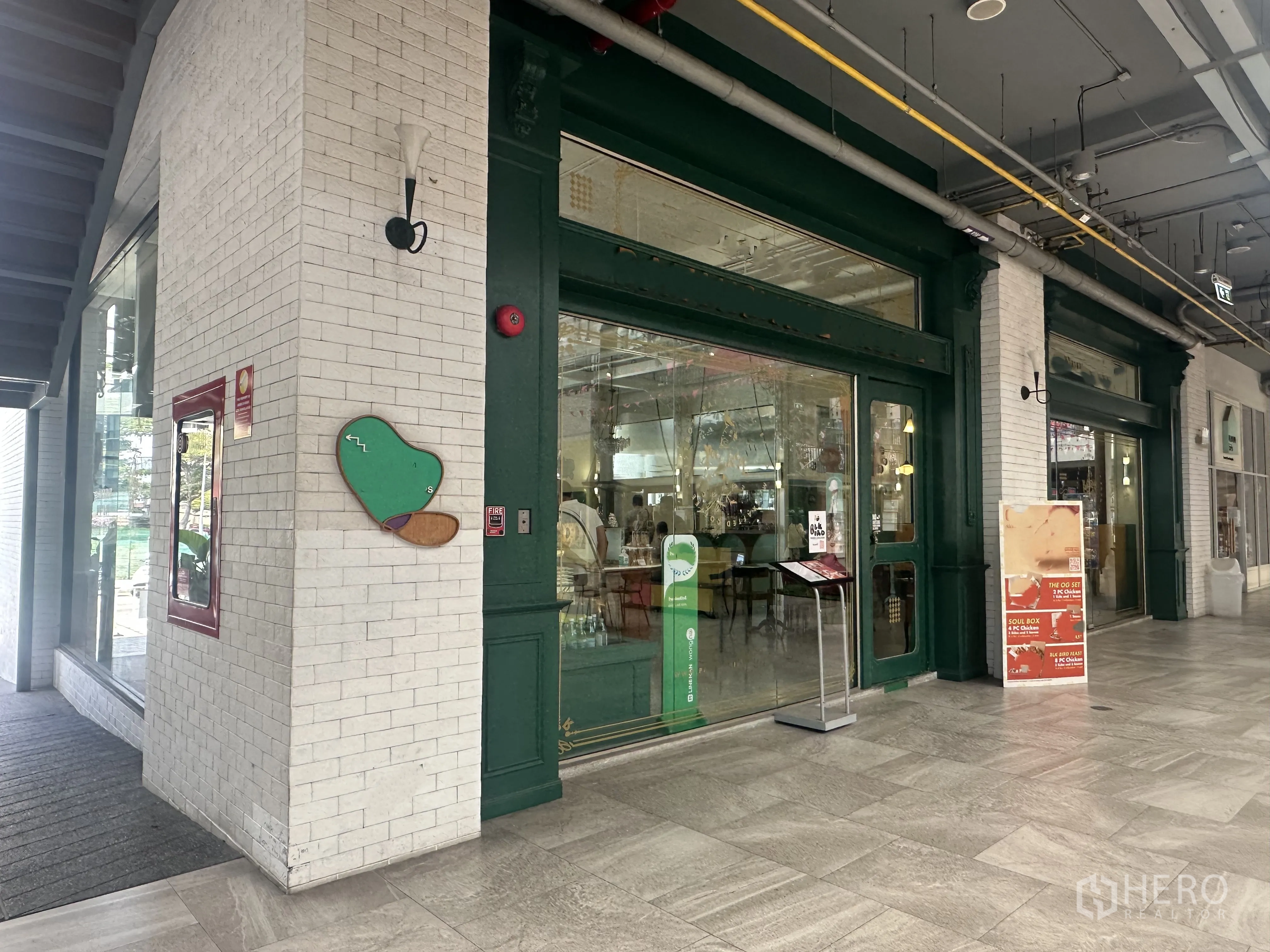 Retail Space for sale in Khlong Toei, Bangkok - Wide retail unit facade along a covered corridor, glass doors with signage, exposed ceiling pipes, and tiled flooring.