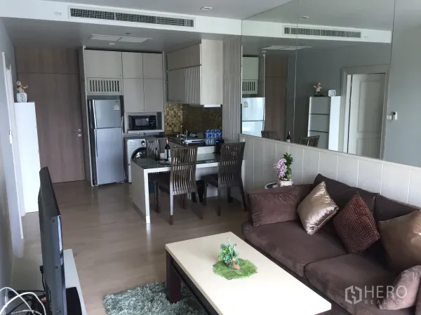 Condominium for rent in Bangkok - Open-plan lounge with mirrored wall, sofa, TV and dining area beside a compact kitchen.