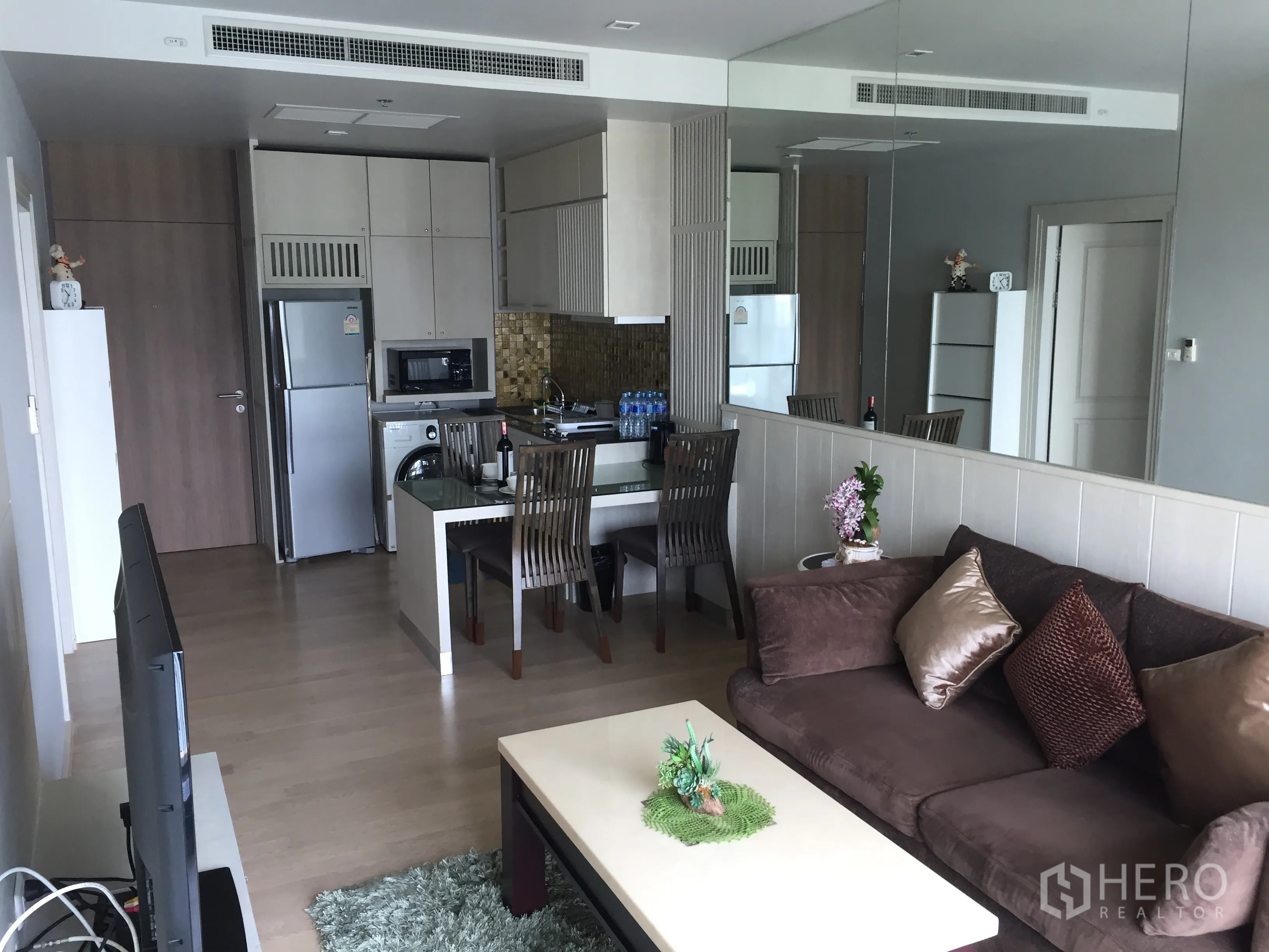 Condominium for rent in Bangkok - Open-plan lounge with mirrored wall, sofa, TV and dining area beside a compact kitchen.