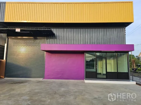 Warehouse for rent in Lam Luk Ka, Pathum Thani - Close front elevation with a large roller shutter beside floor-to-ceiling glass office windows.