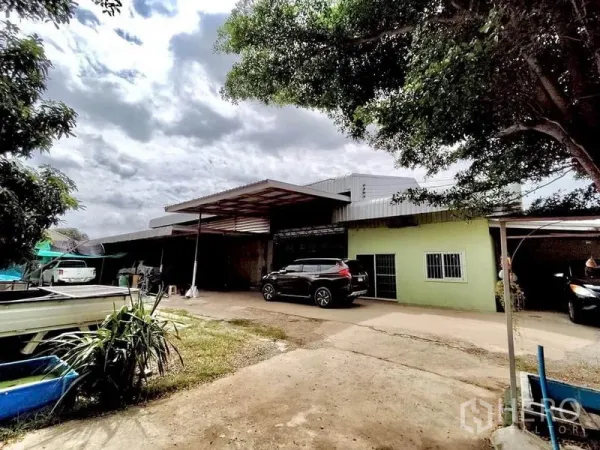 Factory for sale in Lat Lum Kaeo, Pathum Thani - Front view of single-storey factory with covered loading bay, concrete yard and parked vehicles shaded by trees.