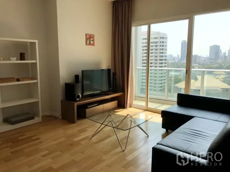 Condominium for rent in Khlong Toei, Bangkok - Bright living room with black leather sofa, glass coffee table, TV console and sliding doors to a balcony with skyline view.