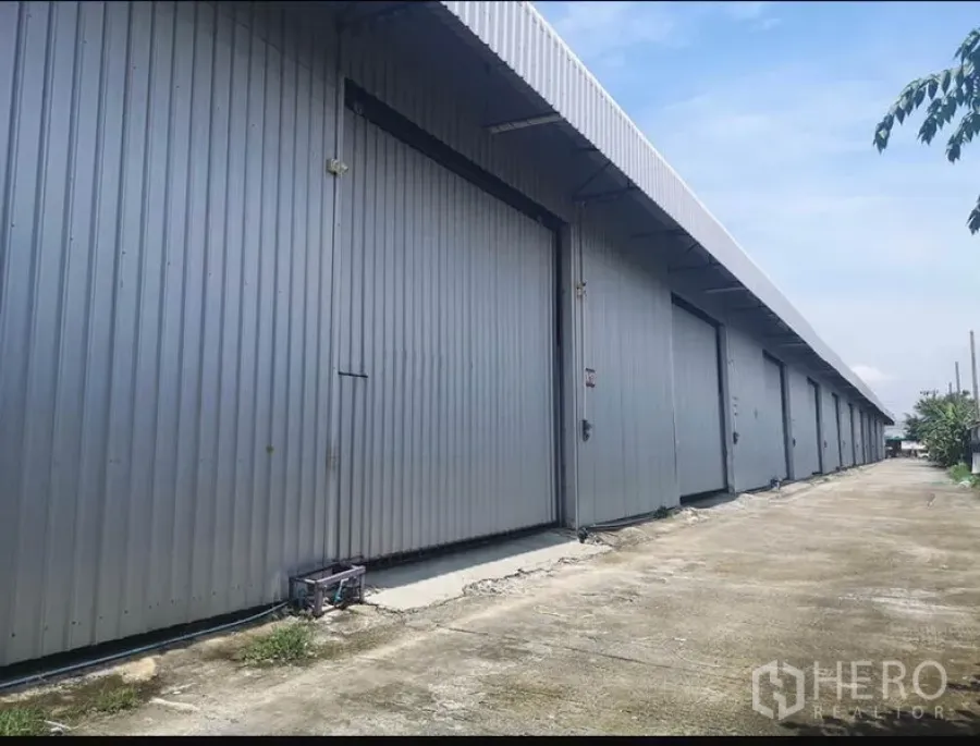 Warehouse for rent in Lam Luk Ka, Pathum Thani - Row of metal-clad warehouse units with large sliding doors and a wide concrete driveway.