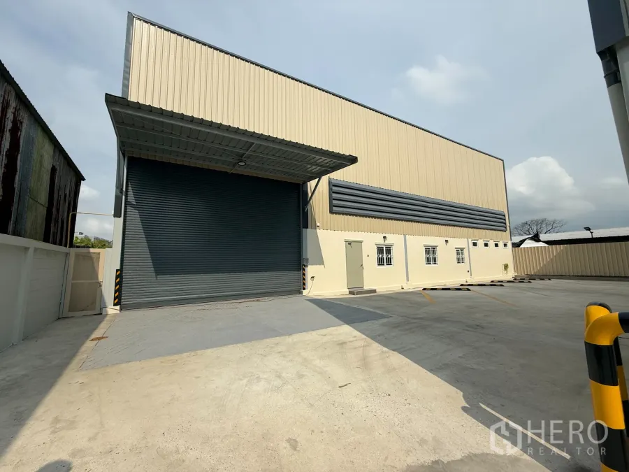 Warehouse for rent in Bang Khen, Bangkok - Modern beige warehouse in Bang Khen, Bangkok with a large roller shutter, covered loading bay, and wide concrete parking yard.
