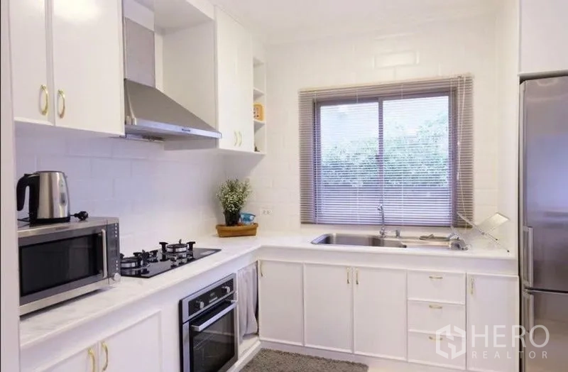 Detached House for rent in Suan Luang, Bangkok - Modern white L-shaped kitchen with gas hob, oven, range hood, sink, and window.