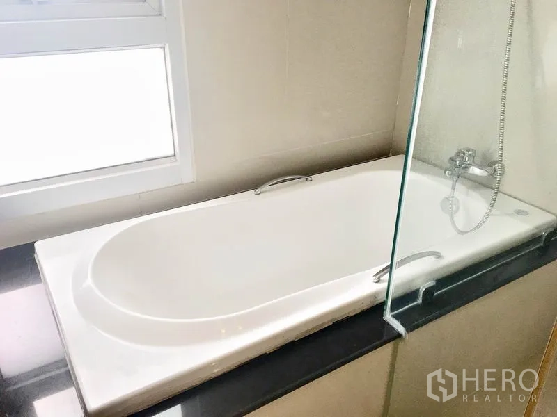 Condominium for rent in Khlong Toei, Bangkok - Close-up of built-in bathtub beside a window with glass partition and shower.