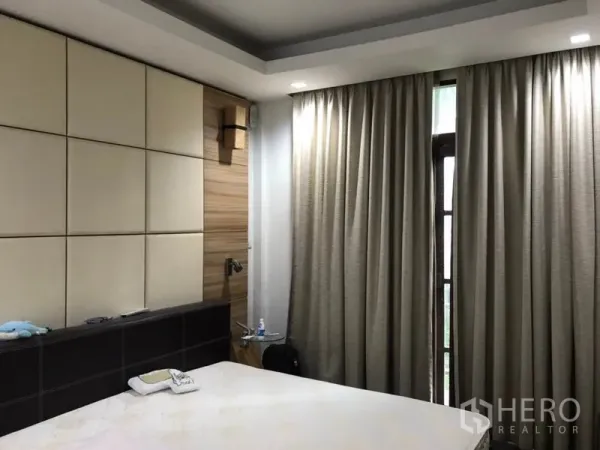 Detached House for sale in Lat Phrao, Bangkok - Primary bedroom with padded headboard wall, wood accents, tray ceiling and full blackout curtains.