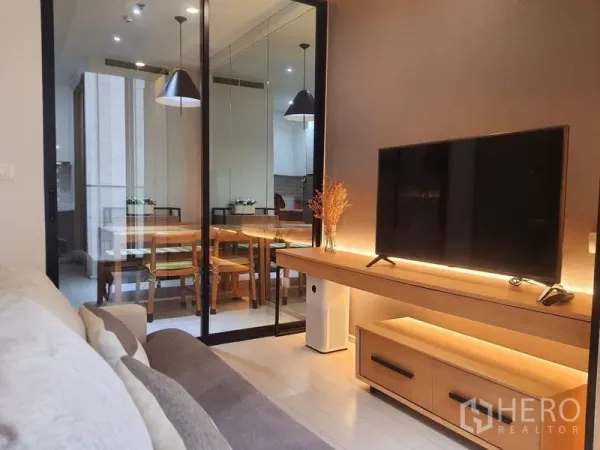 Condominium for rent in Pathum Wan, Bangkok - Cozy living room with TV console and glass partition opening to a dining area with pendant lights.
