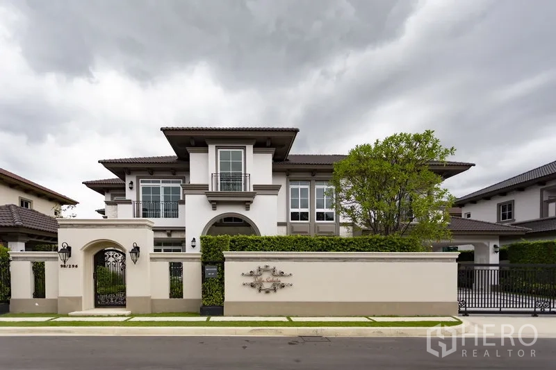 Detached House for rent in Mueang Samut Prakan, Samut Prakan - Front exterior of an elegant two-storey detached house with gated wall, manicured hedge and a tree under a cloudy sky.