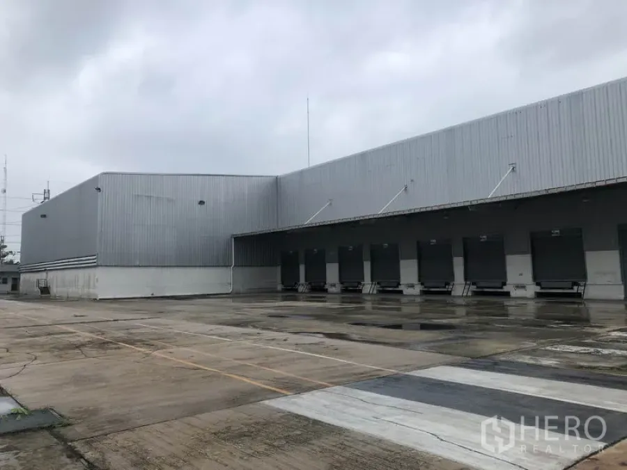 Factory for rent in Phra Nakhon Si Ayutthaya - Large warehouse exterior with multiple dock‑high loading bays and a wide truck yard on a rainy day in Bang Pa‑in.
