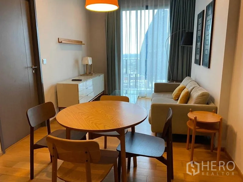 Condominium for rent in Khlong Toei, Bangkok - Open-plan living and dining with round wooden table, sofa and balcony window with sheer curtains.