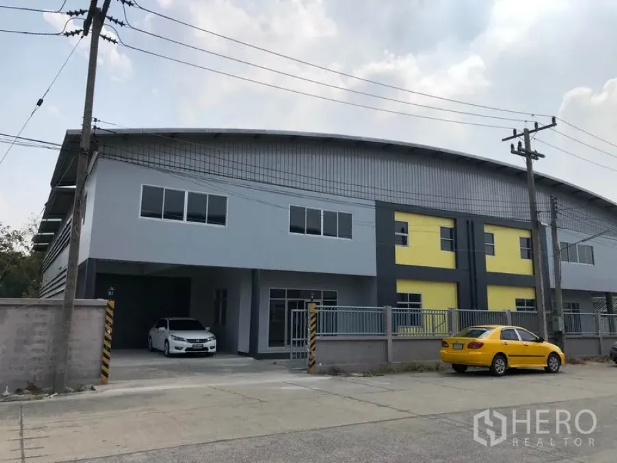 工厂 出租在Lat Lum Kaeo, Pathum Thani - 两层现代工厂外观，灰黄立面，带围栏入口与停车区，临宽阔道路（Lat Lum Kaeo，巴吞他尼）。