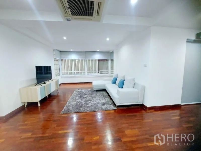 Condominium for rent in Watthana, Bangkok - Living room with L-shaped white sofa, rug, TV unit, ceiling cassette AC and wide windows.