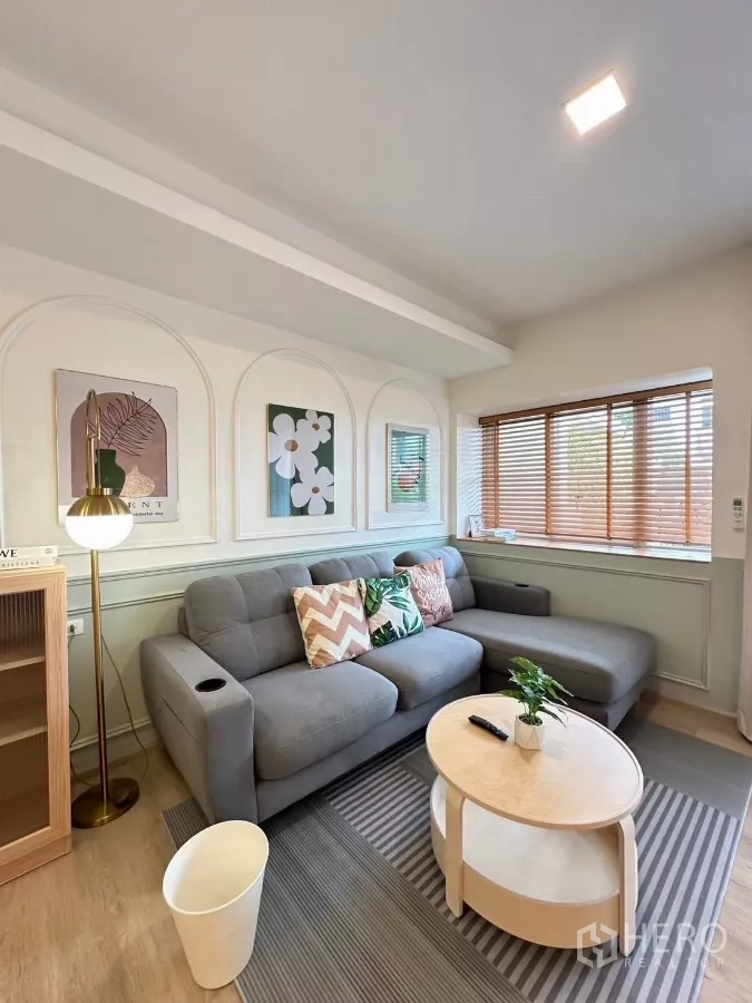 Townhouse for rent in Prawet, Bangkok - Bright living room with gray L‑shaped sofa, round coffee table, wall art and wooden blinds.