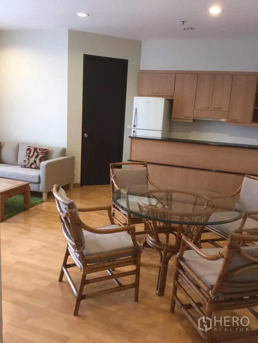 Condominium for rent in Khlong Toei, Bangkok - Open-plan living and dining area with rattan chairs, glass table, sofa and a kitchenette with wood cabinets.