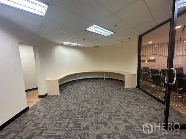 Office Space for rent - Curved workstation area with a long built-in desk, carpet tiles and a glass door to an adjacent meeting room.
