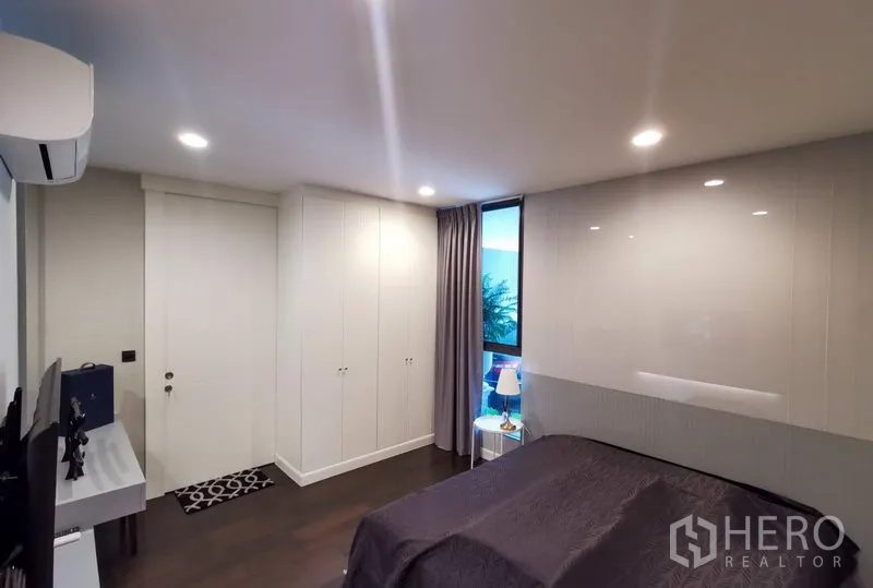 Detached House for rent or sale in Bang Kapi, Bangkok - Calm bedroom with built-in wardrobes, dark wood flooring, and a tall window with curtains.