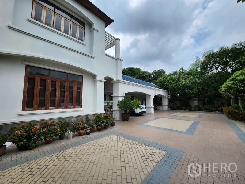 Detached House for rent in Prawet, Bangkok - Wide paved driveway of a white two‑storey detached house with arched carport, potted plants and landscaped edges.