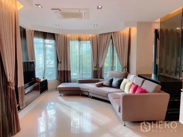 Detached House for rent or sale in Prawet, Bangkok - Corner lounge with bay windows, sheer curtains and an L-shaped sofa facing a TV wall.