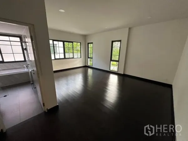 Detached House for rent or sale in Bang Phli, Samut Prakan - Expansive bedroom with dark wood flooring, corner windows and an ensuite bathroom visible through a doorway.