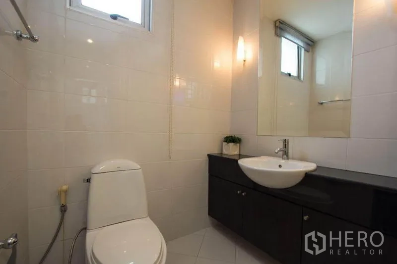 Condominium for rent in Khlong Toei, Bangkok - Compact bathroom with toilet, wall-mounted basin on a dark vanity, large mirror, and window.