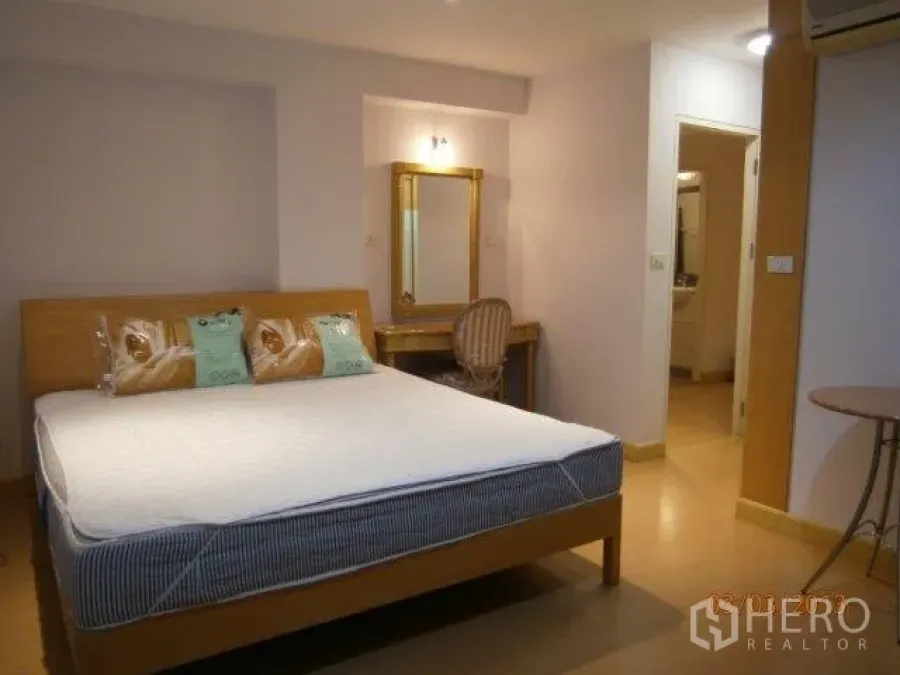 Condominium for rent in Khlong Toei, Bangkok - Bright master bedroom with wooden bed, vanity desk with mirror, and a doorway leading to the bathroom.