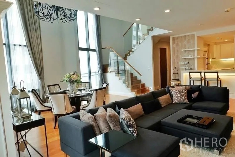 Condominium for rent in Khlong Toei, Bangkok - Luxury condo living area with double-height ceiling, sectional sofa, round dining table, glass staircase and open kitchen with breakfast bar.