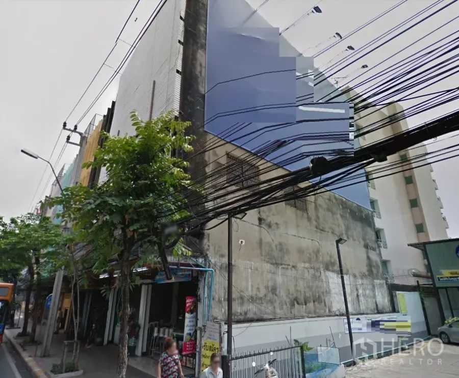 Shophouse for rent in Phra Khanong, Bangkok - Corner exterior of a multi‑storey shophouse block on a Bangkok side street with overhead utility lines and trees.