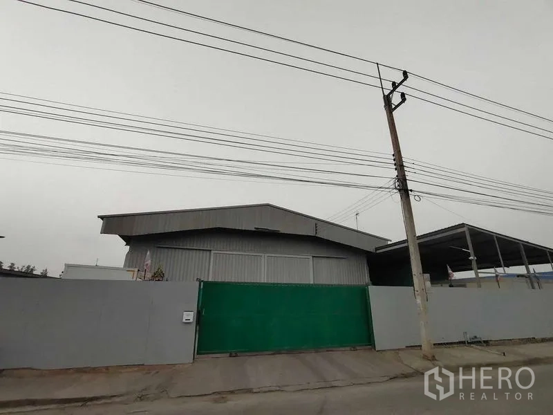Warehouse for sale in Lam Luk Ka, Pathum Thani - Street view of a metal-clad warehouse with a green sliding gate and overhead power lines.