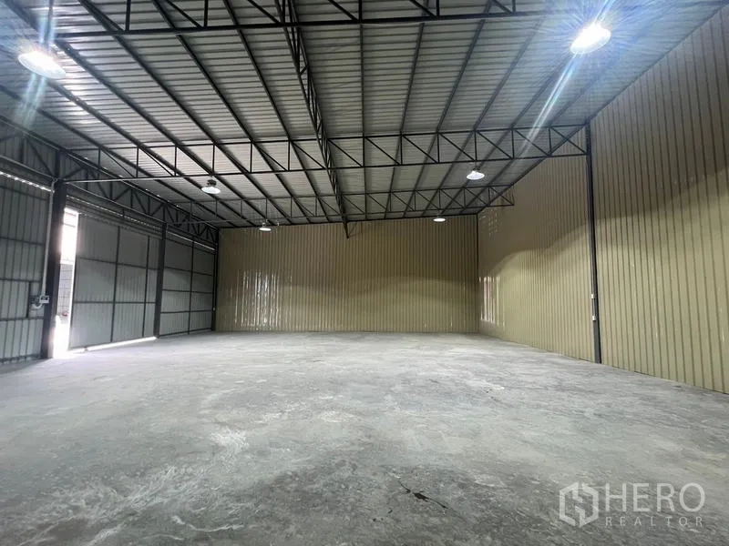Warehouse for rent in Phra Khanong, Bangkok - Angled interior showing beige metal wall, open floor space, and bright overhead lighting.