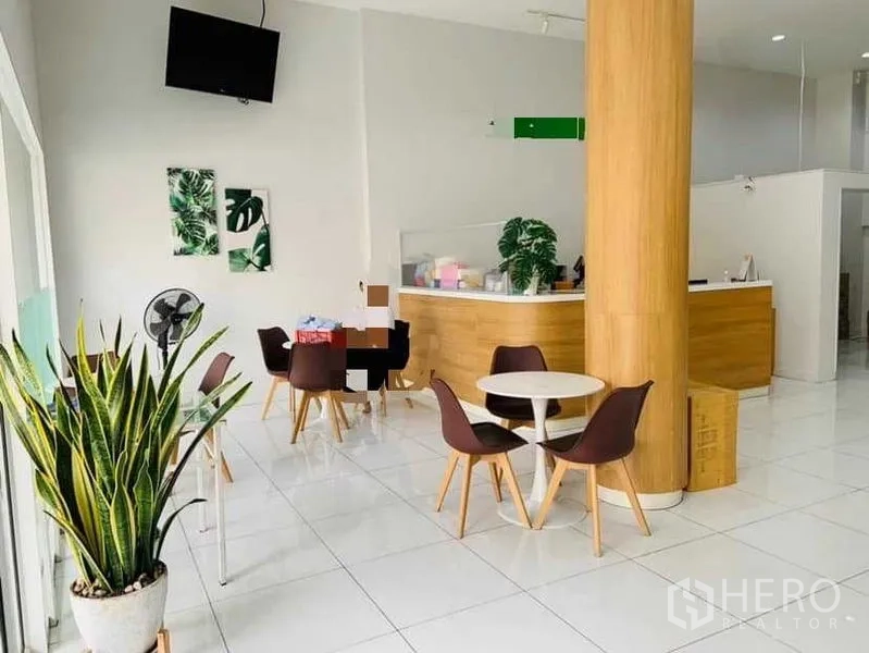 Shophouse for rent or sale in Khlong Toei, Bangkok - Bright ground-floor lobby with wood reception counter, round tables, tall pillar, indoor plants and glossy white tiles.
