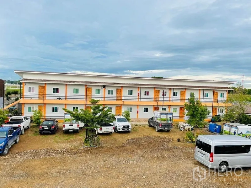 Warehouse for rent or sale in Pathum Thani - Long two-story apartment building with parking area and multiple vehicles.